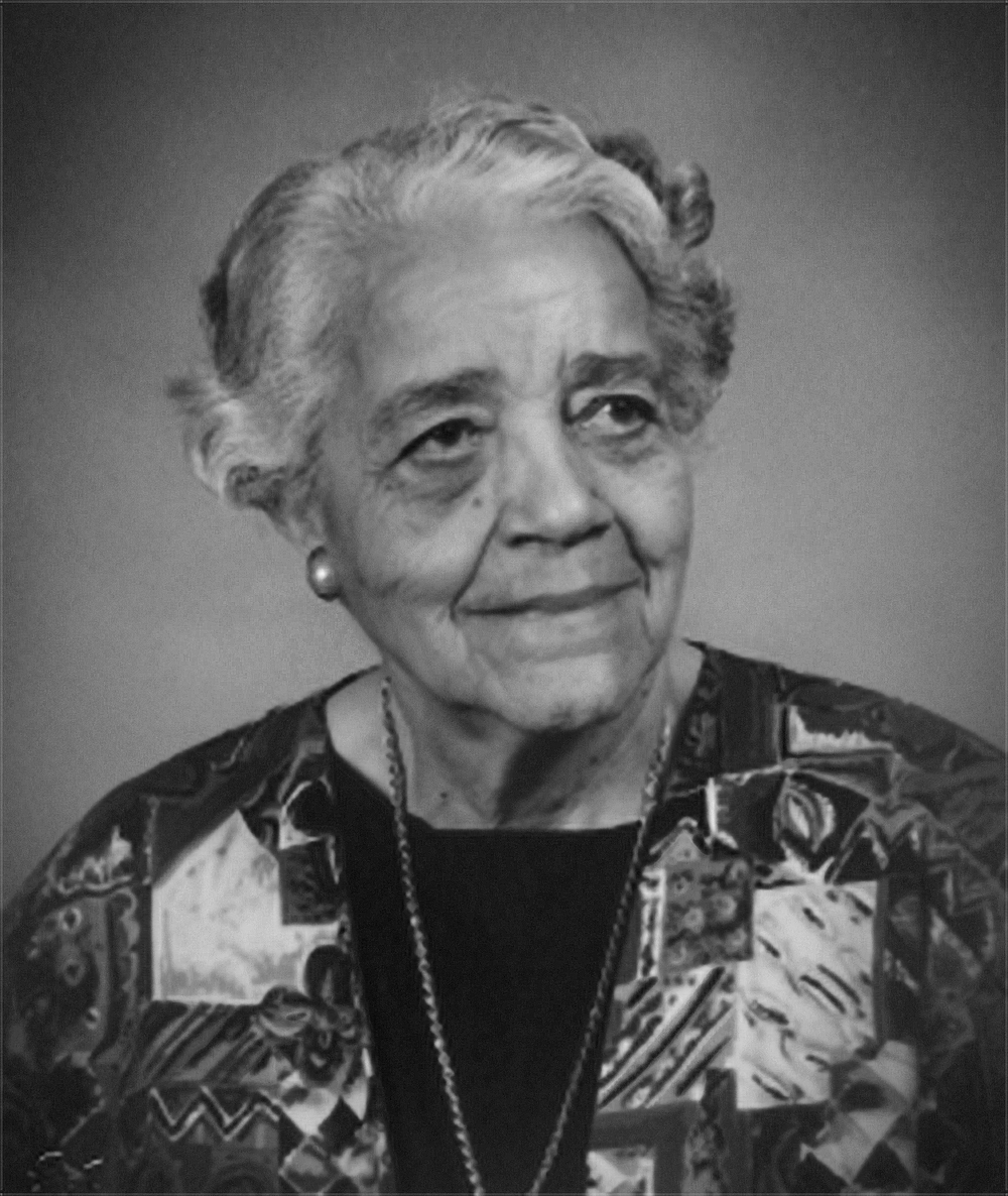 A black-and-white portrait of a woman with short, curly hair. She is wearing a patterned jacket over a dark blouse, accessorized with a necklace and pearl earrings.